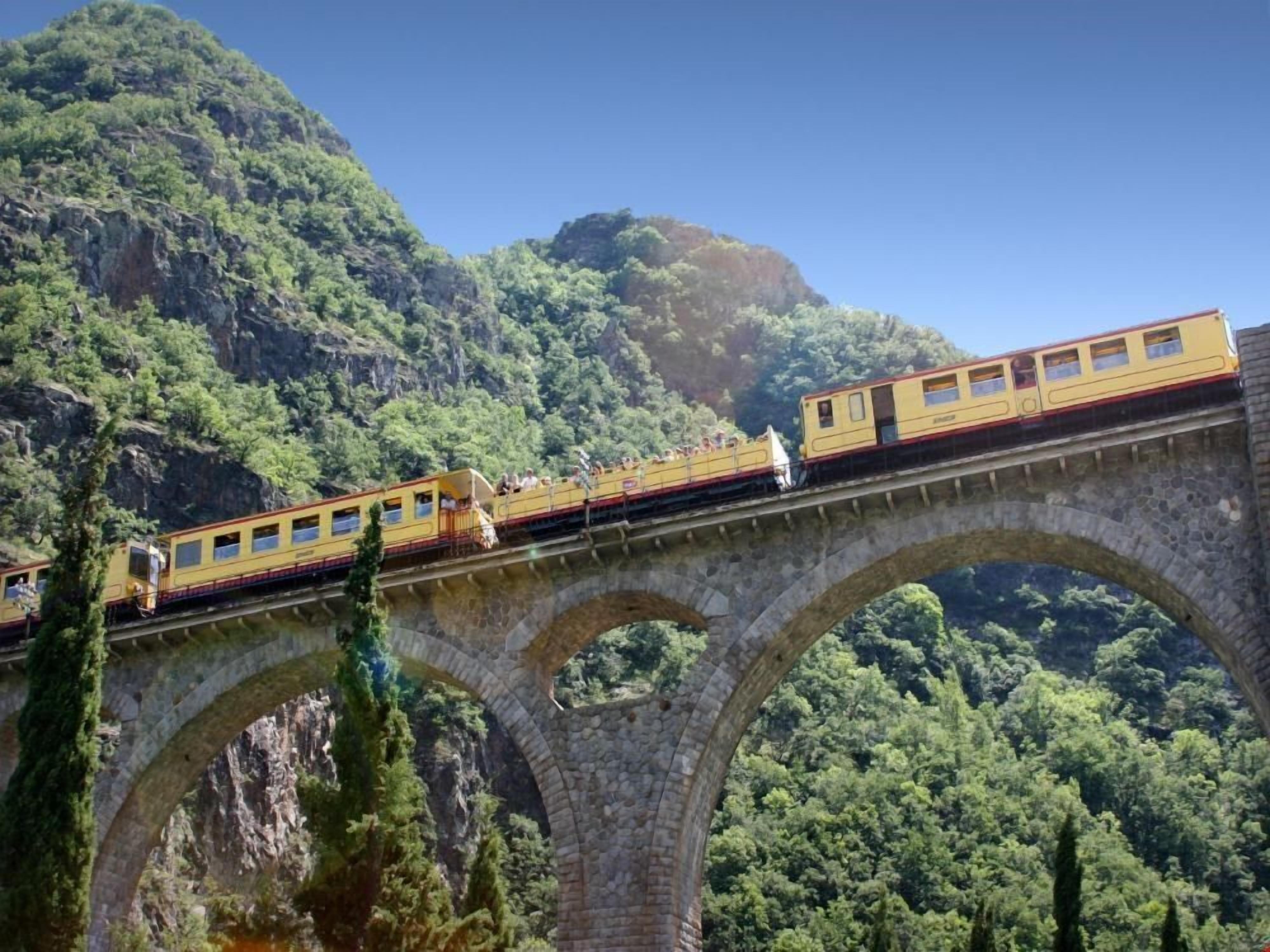 Un tren amarillo sobre un puente de piedra con múltiples arcos, rodeado de un bosque verde y frondoso.