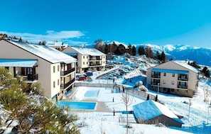 A snow-covered resort with modern buildings, a swimming pool, and a view of snow-capped mountains. - Studio abordable avec cuisinette équipée (Font Romeu)