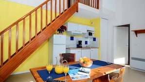 A dining area with a wooden table, chairs, and a fruit bowl. A kitchen with white cabinets and a microwave is visible in the background. - Appartement duplex à quelques pas du centre (Font Romeu)