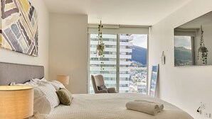 Ein Schlafzimmer mit einem großen Bett, einem Sessel, einem Fenster mit Blick auf Gebäude und einen Berg sowie einer Wand mit einem geometrischen Gemälde.