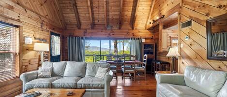 A cabin interior with wooden walls, a sofa, a dining table, and a view of the outdoors.