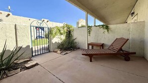 A covered patio area with a wooden lounge chair, a small table, and a gate leading to a street. - Amira Resort #17 | Studio | Near Pool & Pickleball (Saint George)
