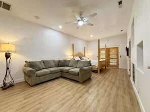 A room with a sectional sofa, a bed with a wooden headboard, a ceiling fan, and a floor lamp. - Amira Resort #17 | Studio | Near Pool & Pickleball (Saint George)