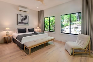 A bedroom with a bed, bench, chair, bedside tables, and a large window with a view of greenery.