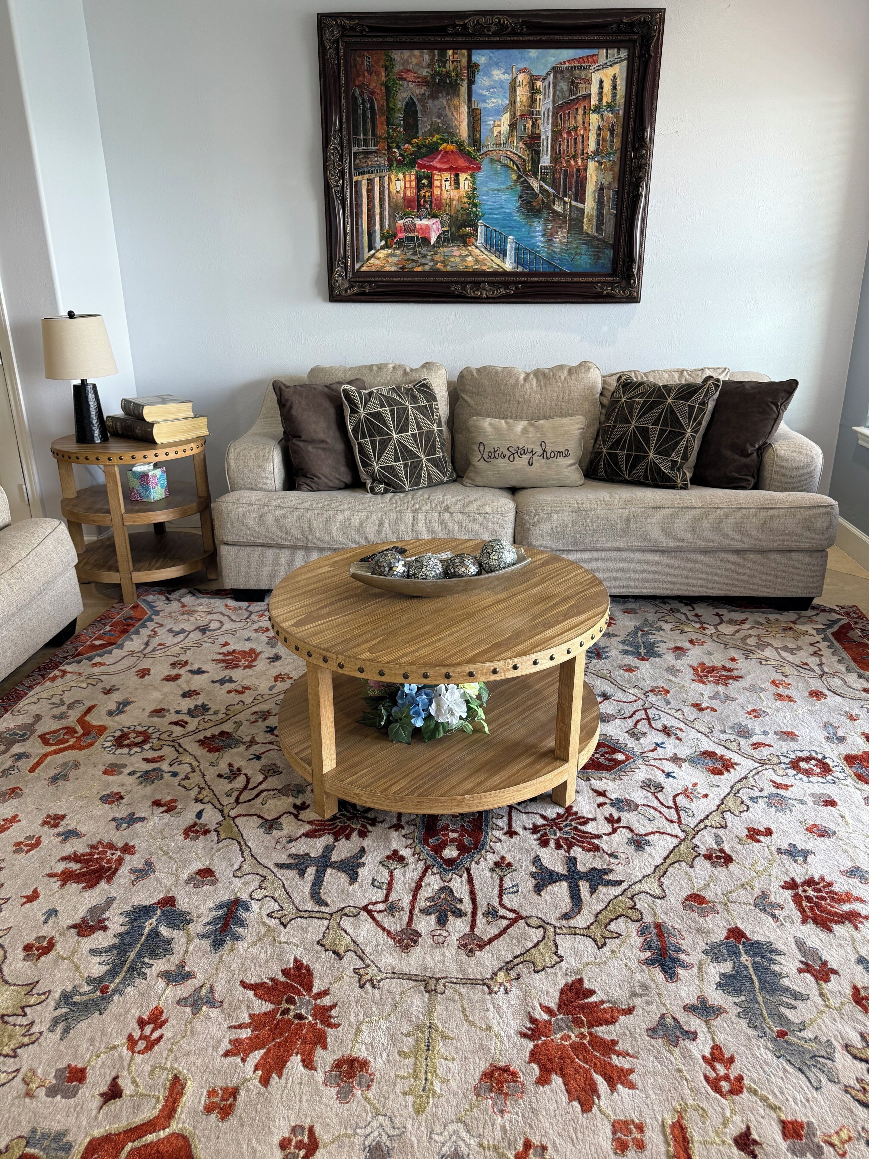 Uma sala de estar com sofá, mesa de centro com objetos decorativos, tapete estampado e um quadro na parede.