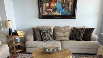 Uma sala de estar com sofá, mesa de centro com objetos decorativos, tapete estampado e um quadro na parede.