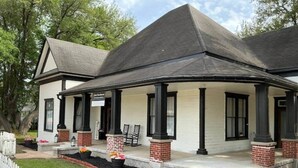 A two-story house with a covered porch, brick pillars, and a white picket fence. - The Anna Bell Suite at historic Jeter Farmhouse (Gladewater)