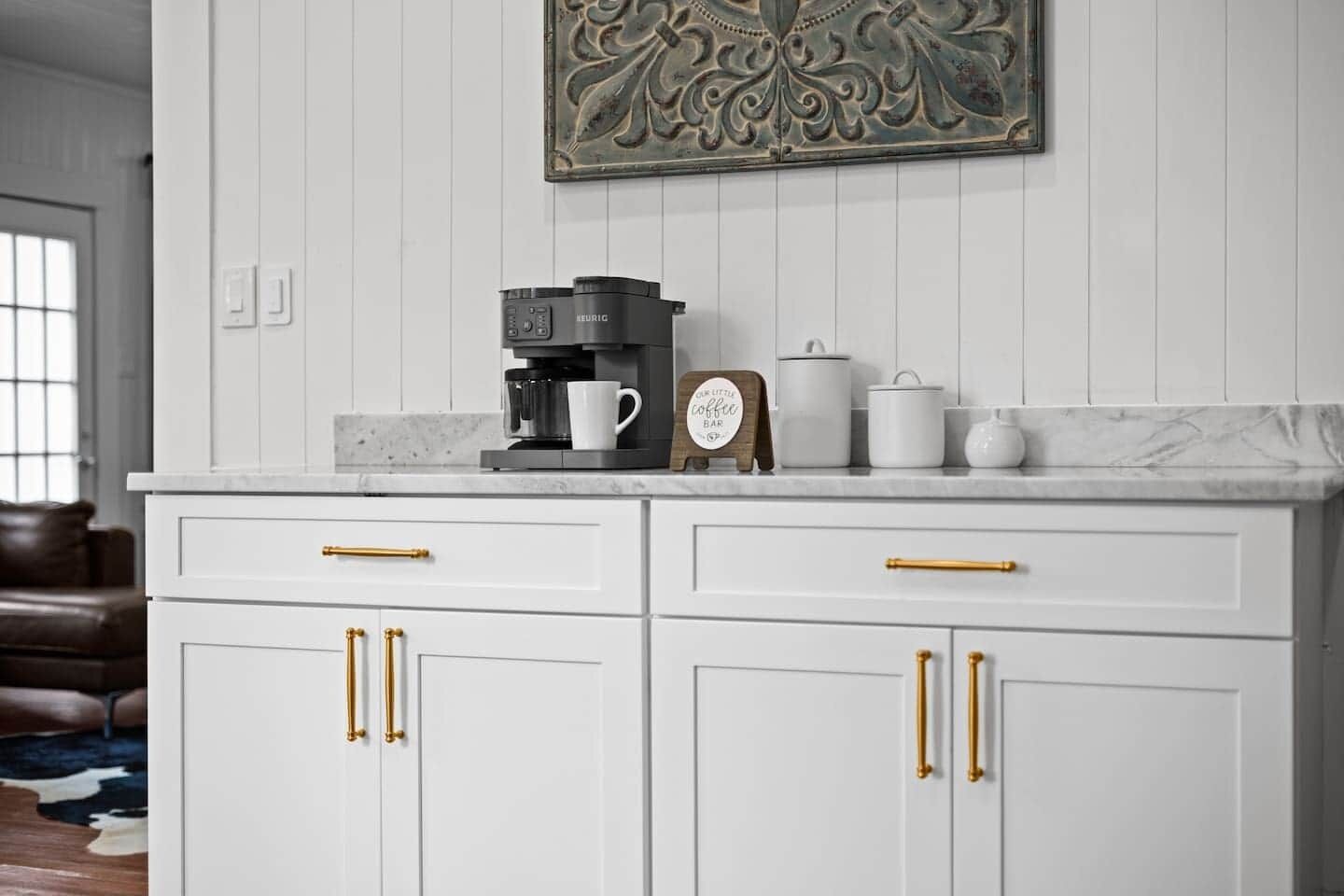 Une cuisine avec des meubles blancs, des poignées dorées, une machine à café, une horloge et un élément décoratif mural.