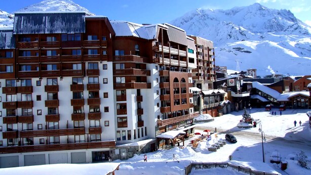 Um hotel com várias andares e varandas, uma estação de esqui ao fundo, e paisagem coberta de neve.