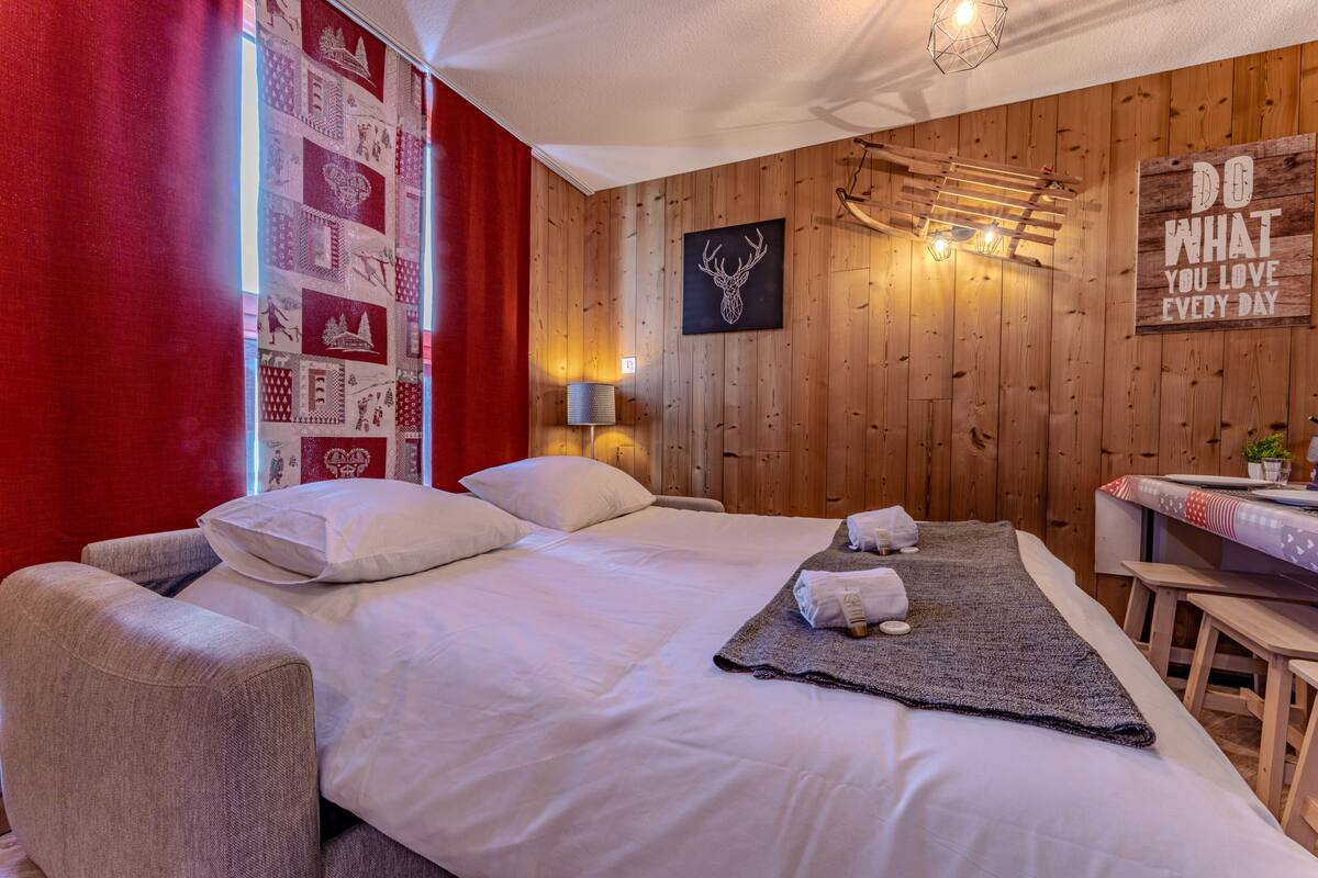 Habitación de hotel con pared de madera, cama con ropa blanca, cortina roja, cuadro de cabeza de ciervo en la pared y un letrero con una frase motivacional.