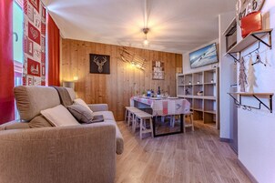 Una sala de estar acogedora con un sofá, una mesa de comedor con bancos, un televisor montado en la pared y una estantería.