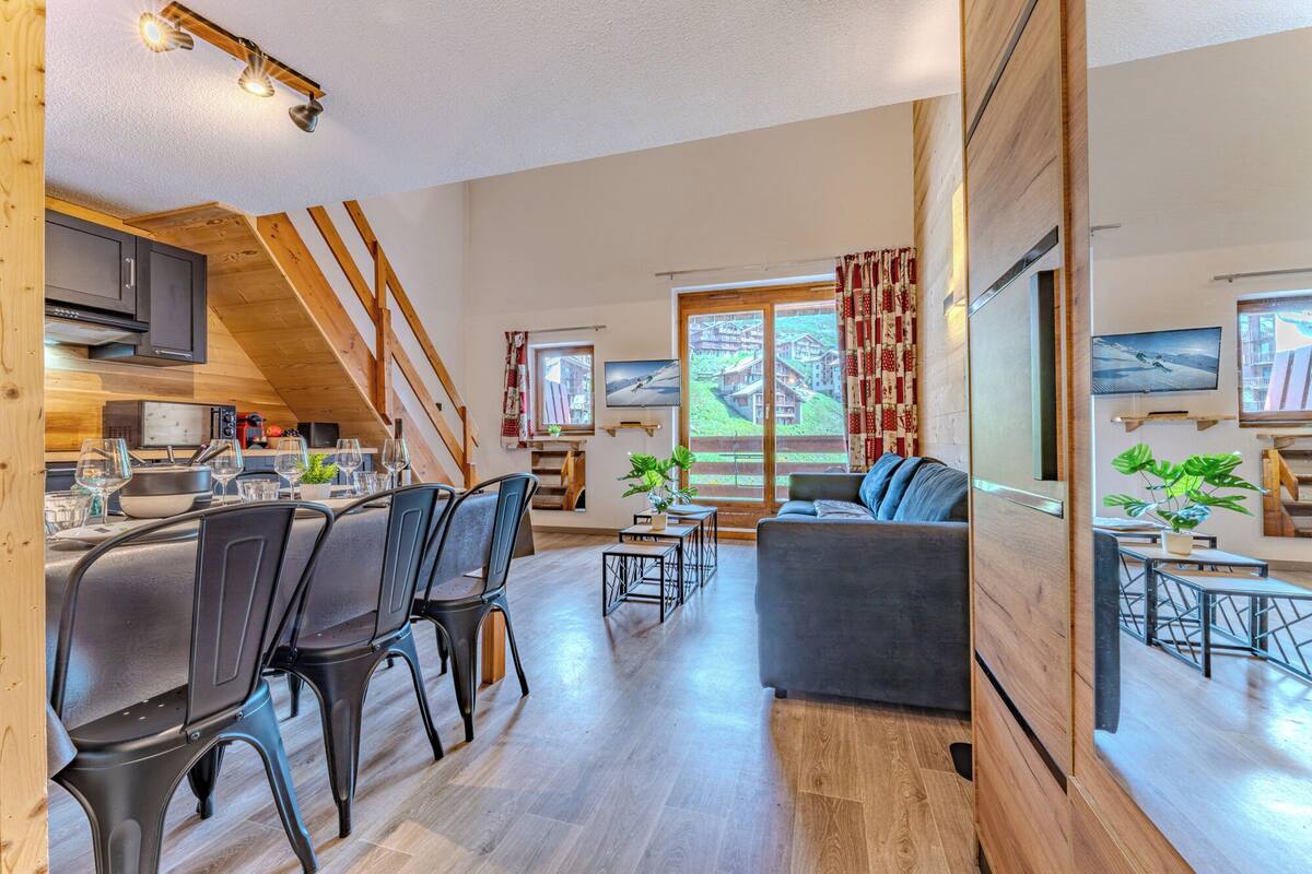 Una sala de estar moderna con un sofá, comedor con sillas, un televisor y una cocina con vigas de madera.