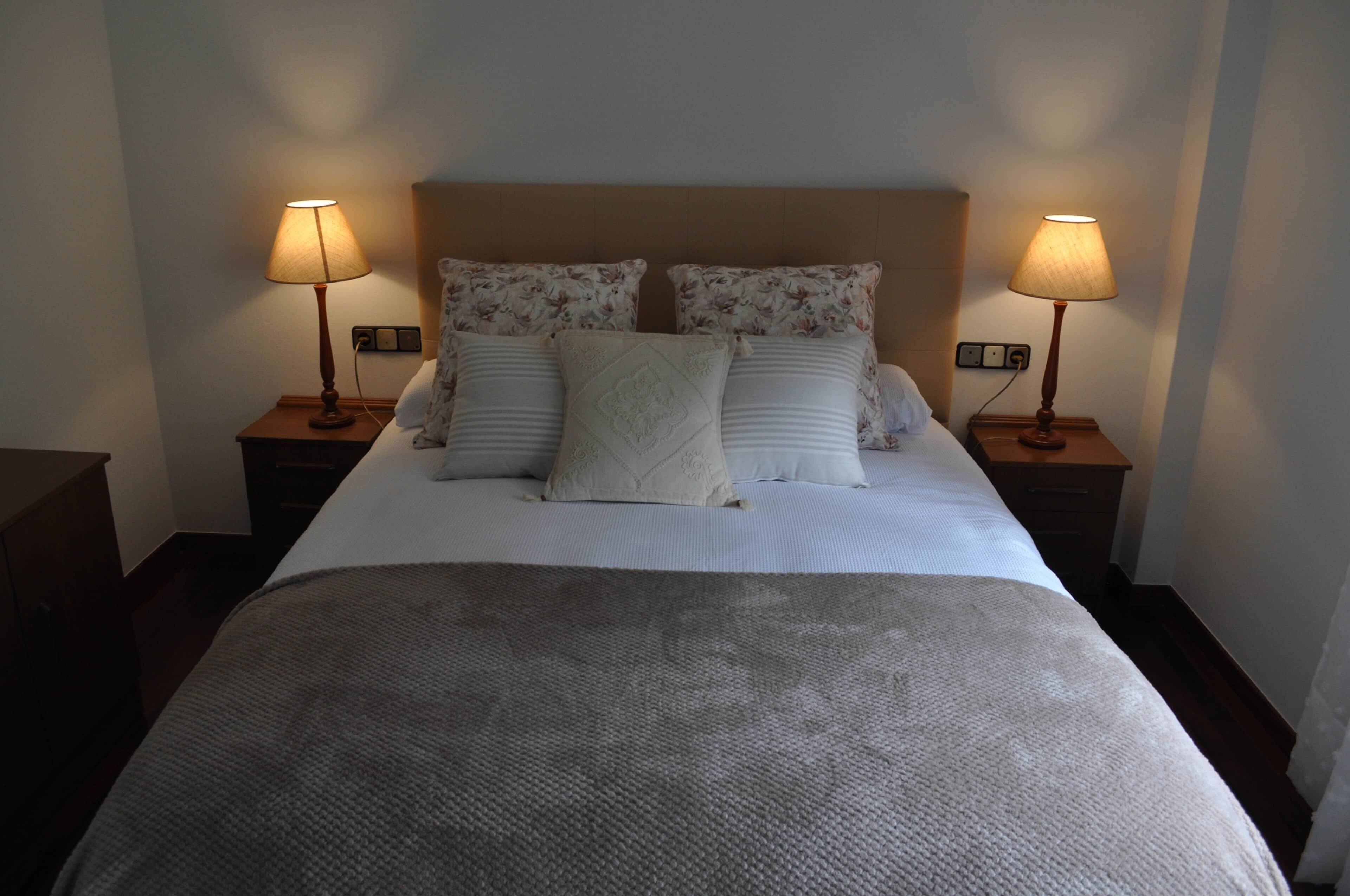 Ein ordentlich bezogenes Bett mit grauer Decke, zwei Nachttischlampen und Kissen mit Blumenmuster.