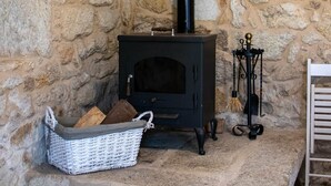 Un mur de pedra, un forn de llenya negre, una cistella de vimet amb llenya i un suport metàl·lic amb una escoba.