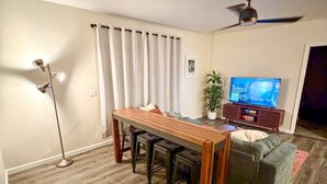 Ein Wohnzimmer mit einem Holzesstisch, vier Barhocker, einem grünen Sofa, einem Fernseher, einem Deckenventilator und einer Stehlampe.