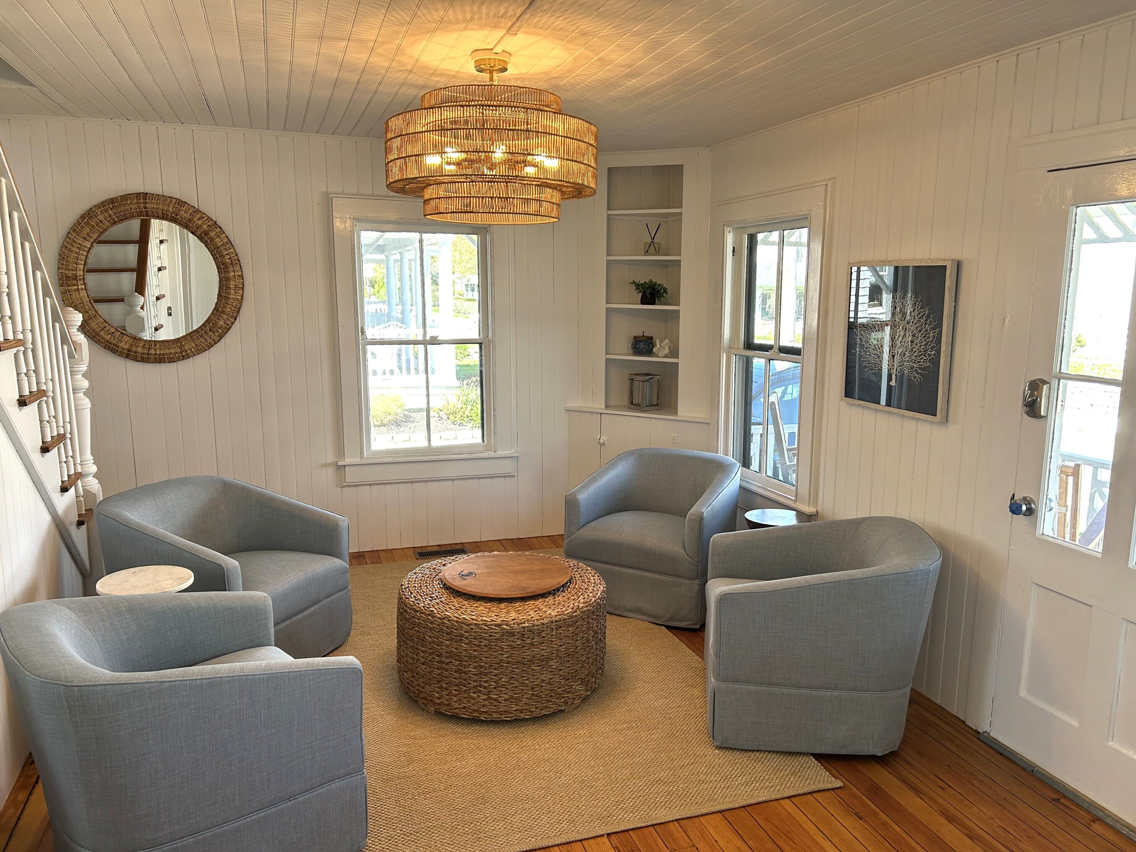 A room with a staircase, two grey armchairs, a round woven ottoman, a large mirror, and a hanging light fixture.