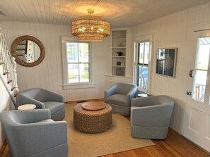 A room with a staircase, two grey armchairs, a round woven ottoman, a large mirror, and a hanging light fixture.