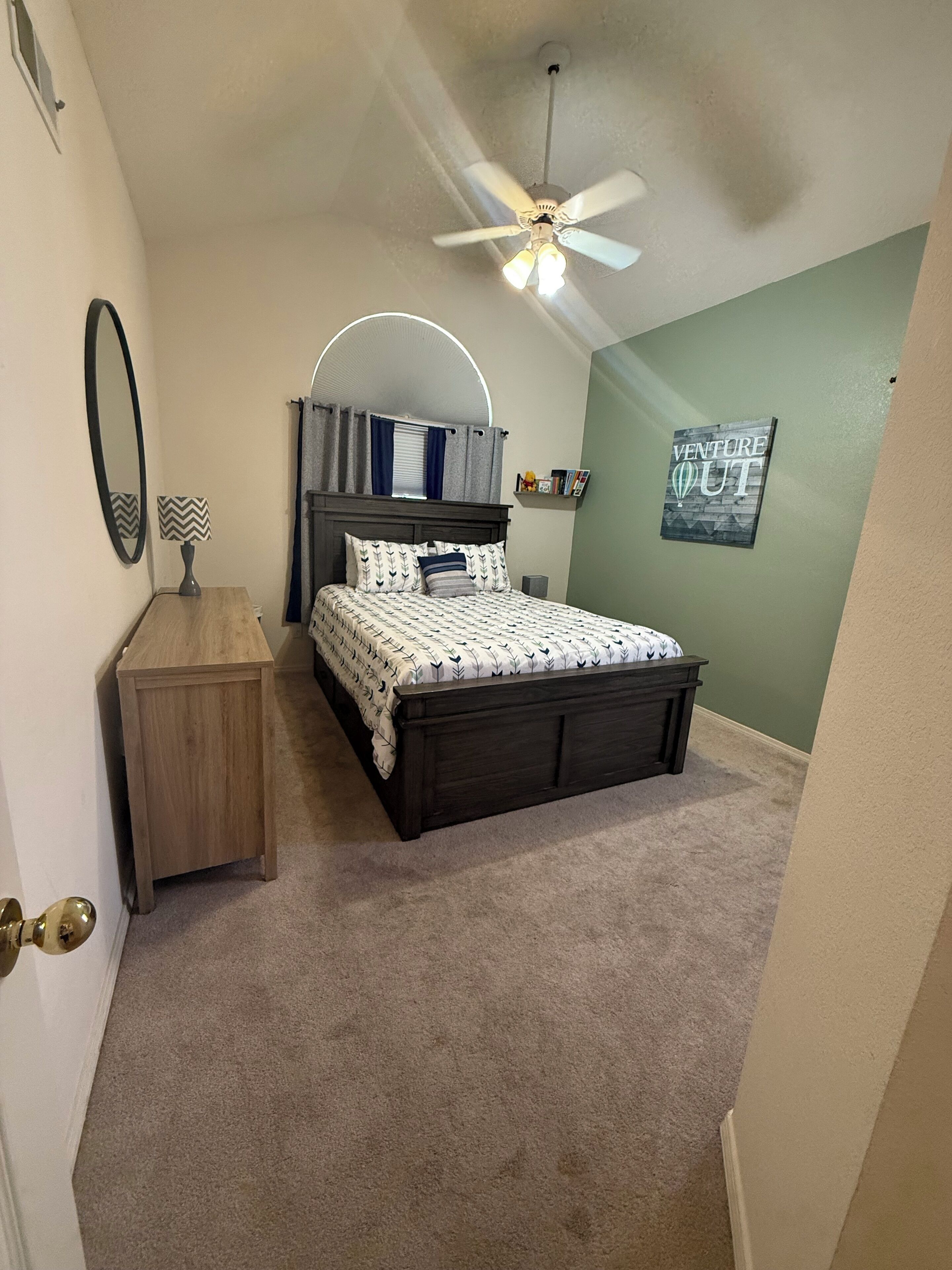 A bedroom with a bed, a nightstand, a mirror, a ceiling fan, and a wall with a 'Venture Out' sign.