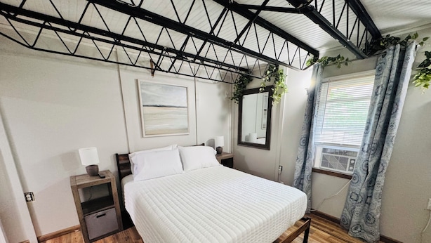 A bedroom with a bed, bedside tables, a mirror, a window with curtains, and a ceiling with exposed beams. - Stylish 4BR Loft • Sleeps 8 • Prime Location (Oswego)