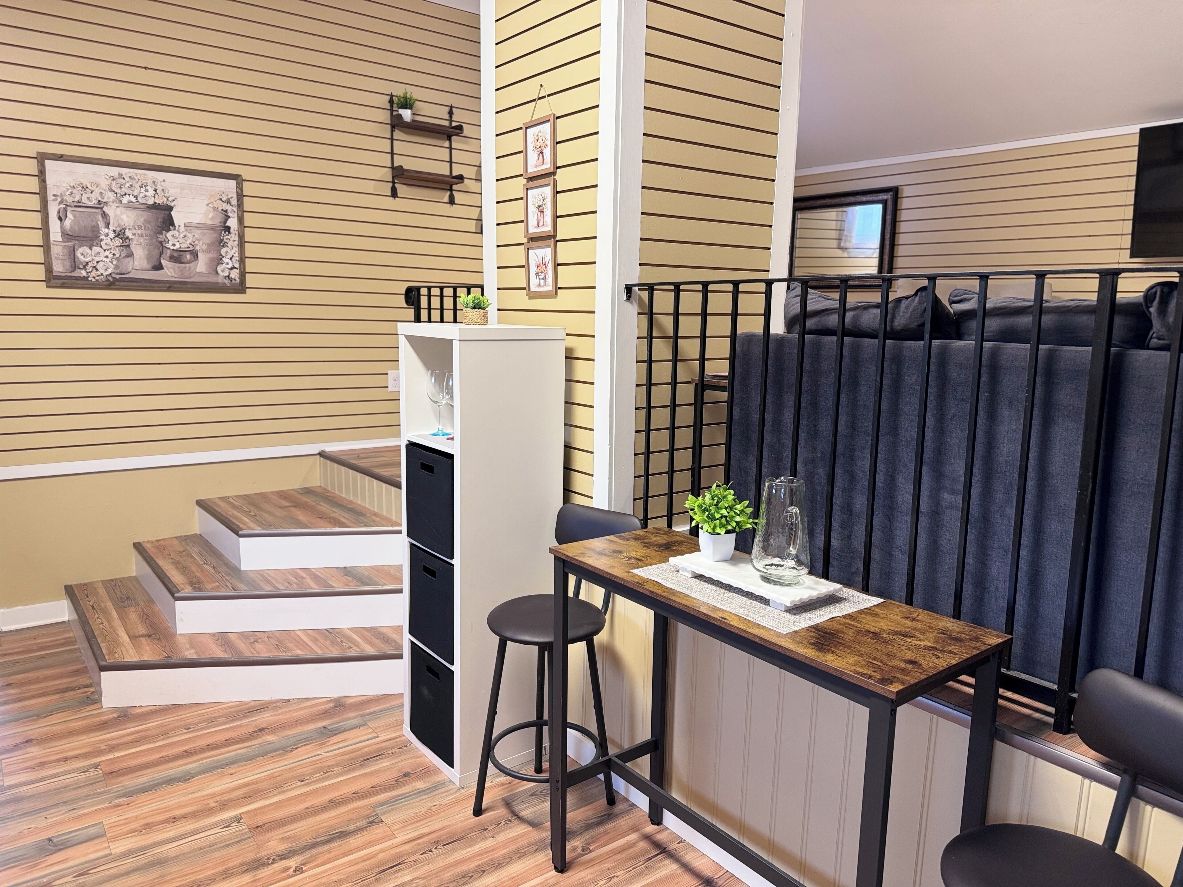Un espace de vie compact doté d’un plancher en bois, d’un petit coin repas avec des tabourets, d’un comptoir de cuisine et d’un escalier menant à un niveau supérieur.