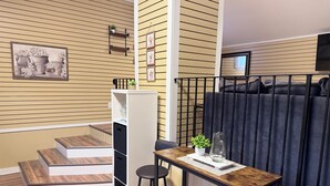 A compact living space with a wooden floor, a small dining area with stools, a kitchen counter, and a staircase leading to an upper level. - Stylish 4BR Loft • Sleeps 8 • Prime Location (Oswego)