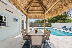 Eine überdachte Terrasse mit Pool, einem Esstisch mit Stühlen und Blick auf Pool und Grünflächen.