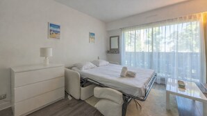 A hotel room with a bed, a bedside table, a chair, a dresser, and a window with curtains. - Studio des Ouvres - Welkeys (Deauville)