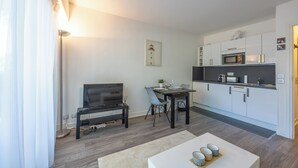 A modern living room with a flat-screen TV, a dining table with chairs, a kitchen area with white cabinets and appliances, and a large window with sheer curtains. - Studio des Ouvres - Welkeys (Deauville)