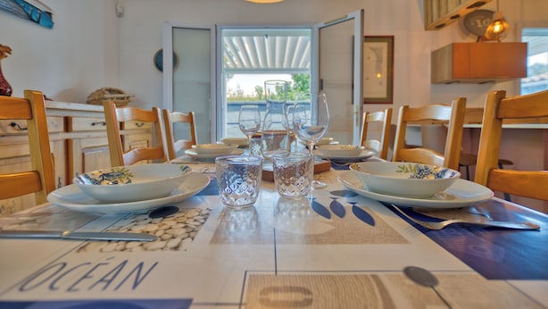 Une table à manger dressée avec des assiettes, des bols, des verres et des couverts, donnant sur l’extérieur grâce à une porte ouverte.