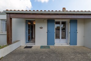 A building with a tiled roof, a door with blue shutters, and a small porch area. - Maison Oléron - Welkeys (Dolus-d'Oléron)
