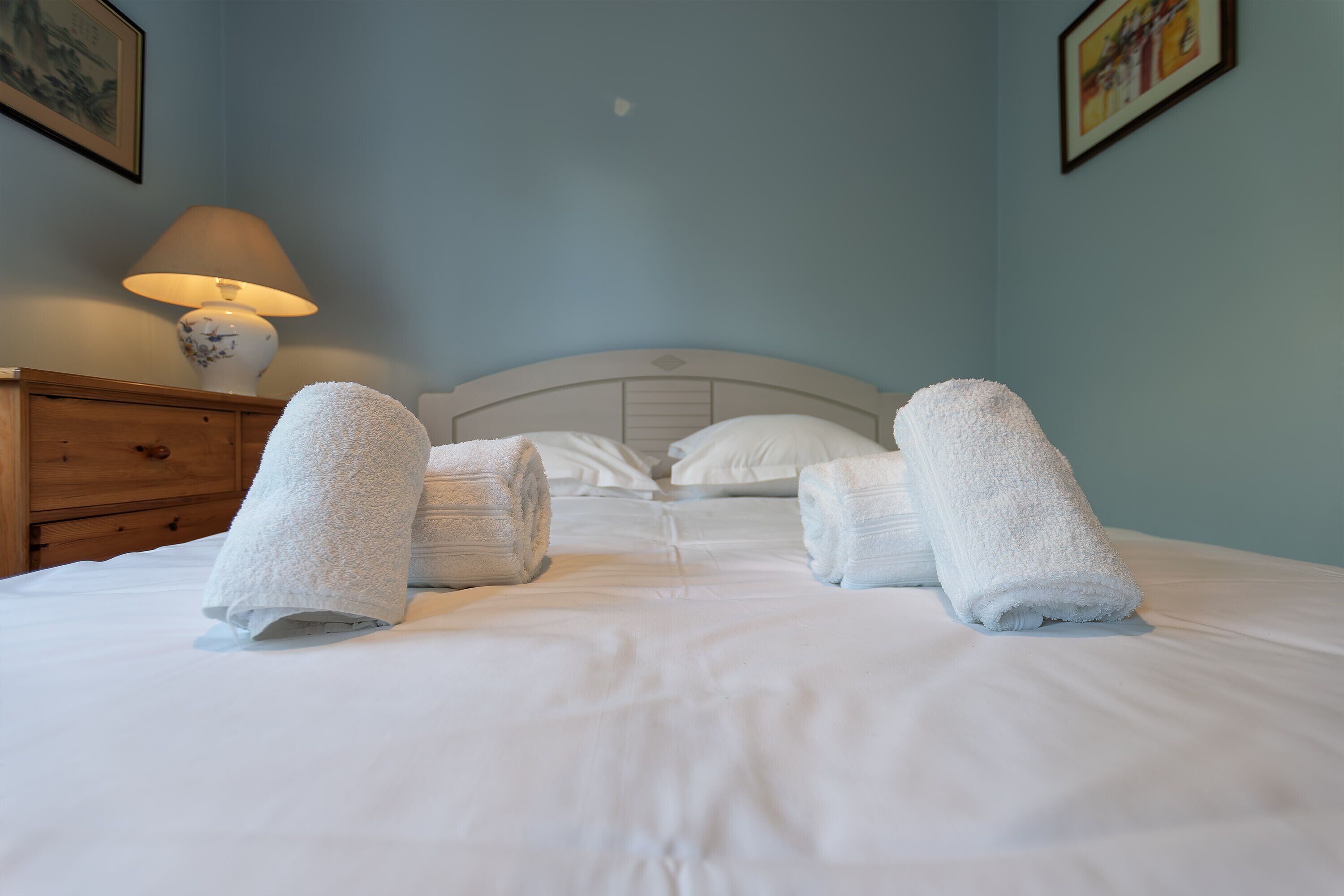Cama bem arrumada com lençóis e toalhas brancas, mesa de cabeceira de madeira com abajur e cabeceira em parede azul claro.