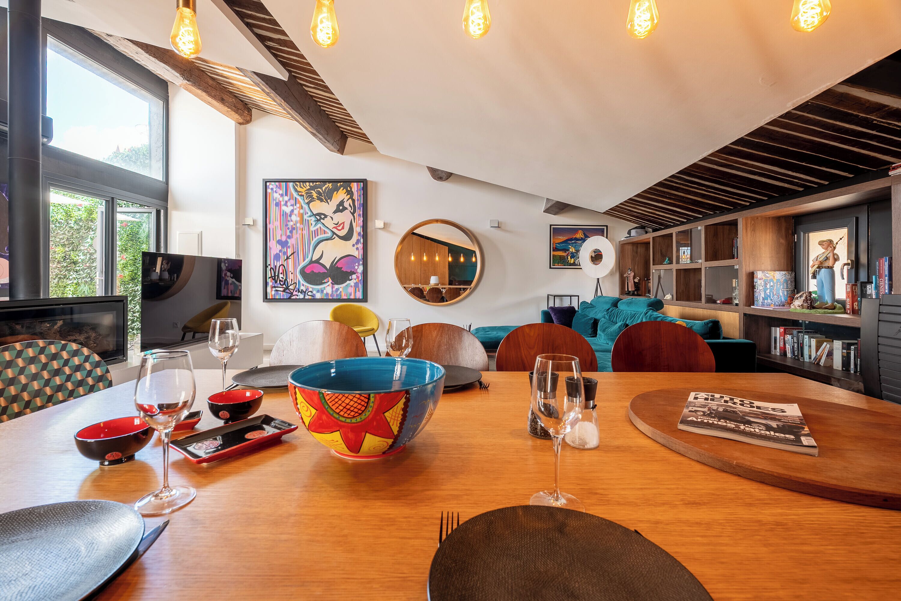 A modern living room with a wooden dining table, colorful bowls, wine glasses, and a bookshelf.
