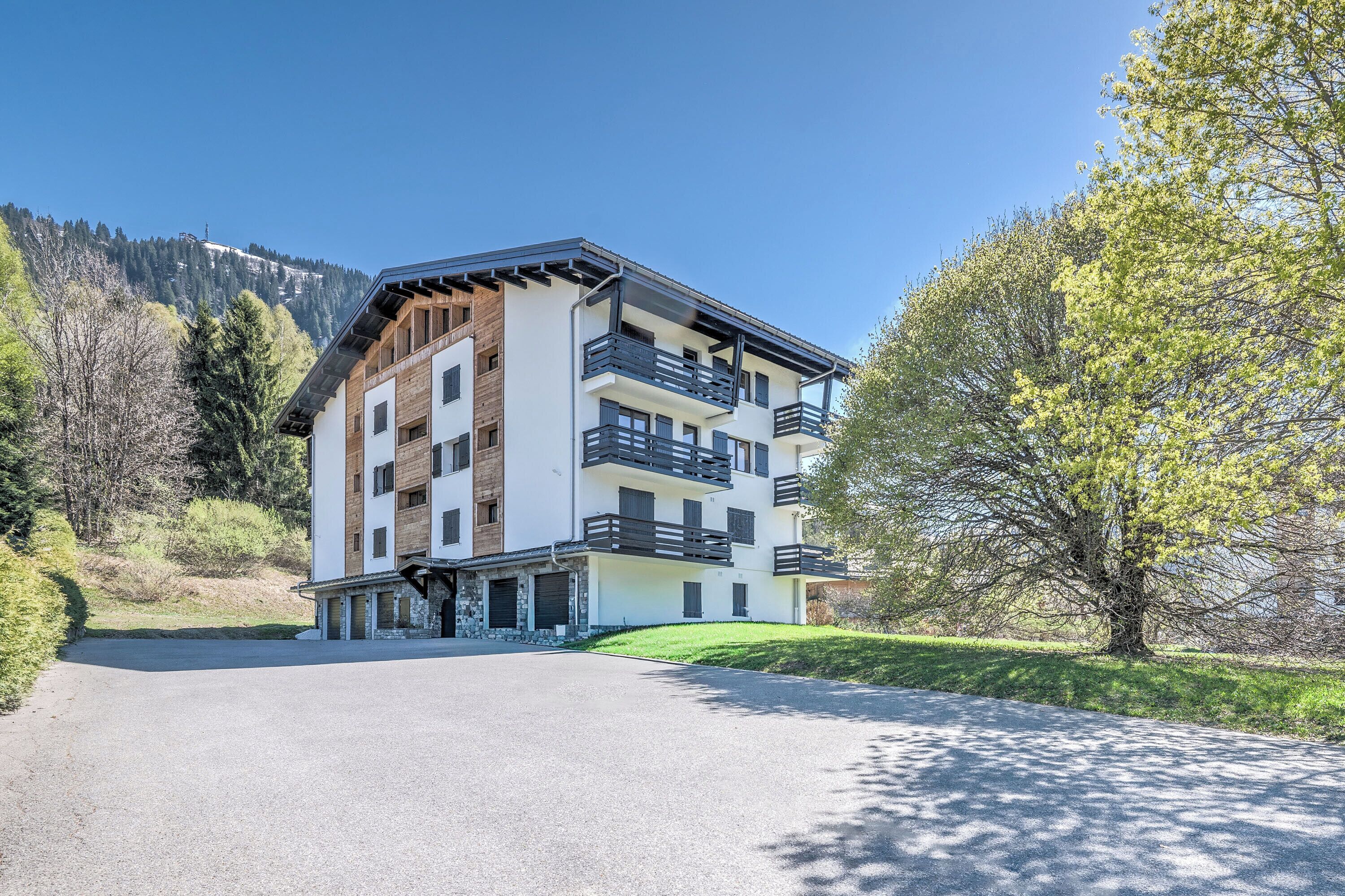 A modern multi-story building with balconies, a driveway, and a mountainous backdrop.