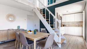 Zona pranzo con tavolo e sedie in legno, una scala e angolo cottura con mobili bianchi.