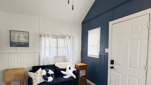 Una habitación con una pared de acento azul oscuro, una puerta blanca, una mesita de noche de madera, un sofá azul con cojines blancos y una mesa redonda de madera, además de un ventilador de techo.