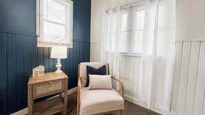 Una habitación con pared azul oscuro, una mesita de noche de madera con una lámpara, una silla con cojín y una ventana con cortinas blancas.