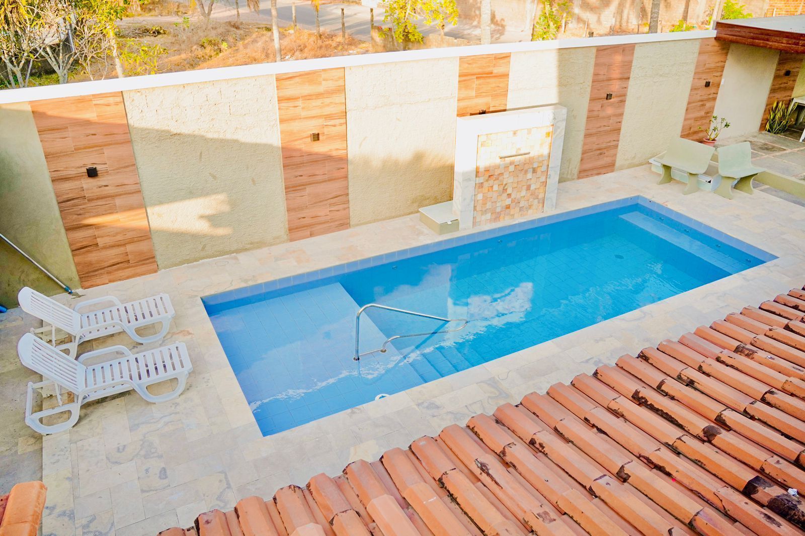 Una piscina rectangular amb una escala metàl·lica, envoltada per un solàrium amb rajoles. Hi ha dues cadiretes blanques i una taula petita amb una cadira.