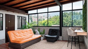 A modern living room with a sofa, a chair, a small table, and a large window offering a view of greenery and buildings. - 302-3 habitacion con baño privado en Coliving con terraza (Medellín)