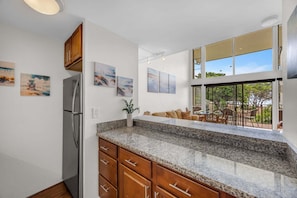 A kitchen with granite countertops, wooden cabinets, and a refrigerator. There are multiple paintings on the walls, and a dining area with outdoor seating visible through large windows. - Maalaea Oceanview 1BD + Loft Getaway (Wailuku)