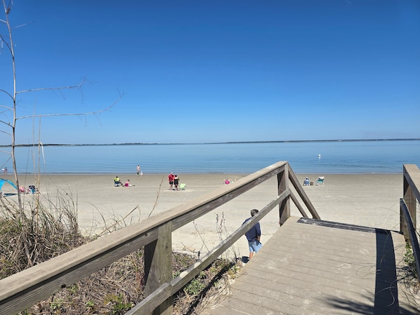 Ein hölzerner Steg führt zu einem Sandstrand mit klarem, blauem Wasser.