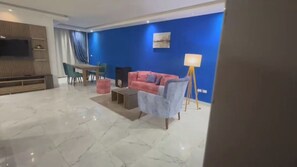 Un salon moderne avec un mur d’accent bleu, un téléviseur à écran plat, un canapé rose, un fauteuil gris et une lampe sur pied.