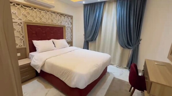 A neatly made bed with a red headboard, a wooden nightstand, a desk with a chair, and a window with blue curtains.