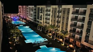Un hotel con una piscina grande, varios balcones y una zona de piscina bien iluminada por la noche.