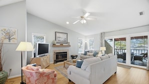 Una sala de estar con sofá, sillón, televisor, chimenea y ventanas amplias.