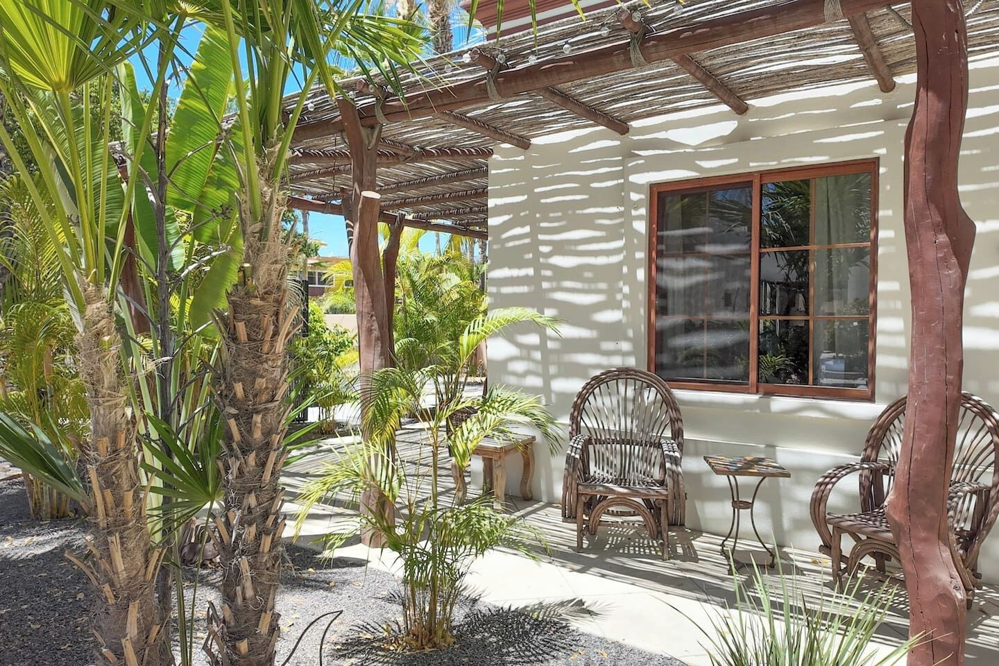 Un edificio blanco con una pérgola de madera, dos sillas de mimbre y una mesa.