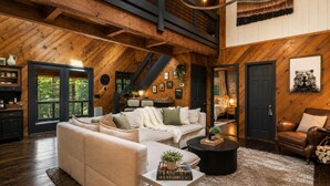A spacious living room with a high ceiling, wooden beams, a large sofa set, a coffee table, and a fireplace. - Cabin Sleeps 12: Screened Hot Tub, Arcade, Dogs (Blue Ridge)