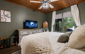 Ein Schlafzimmer mit Bett, Deckenventilator, Fernseher und einem Fenster mit Vorhängen.