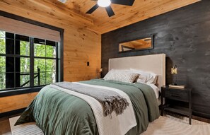 A bedroom with a large bed, a nightstand, a mirror, and a window with a view of trees. - Blue Ridge Cabin Sleeps 12: Games, Views, Hot Tub (Blue Ridge)