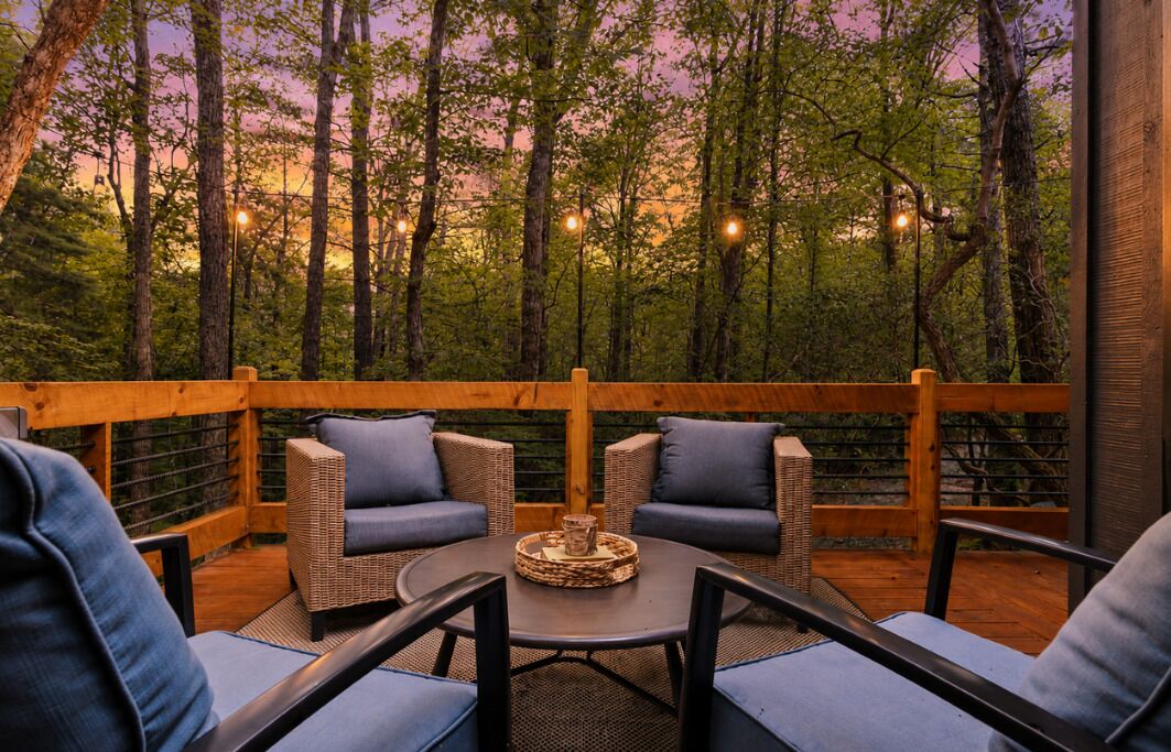 A wooden deck with wicker furniture and a round table, surrounded by trees and string lights.