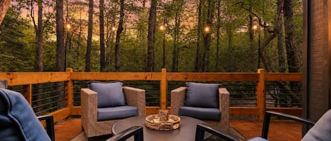 A wooden deck with wicker furniture and a round table, surrounded by trees and string lights.